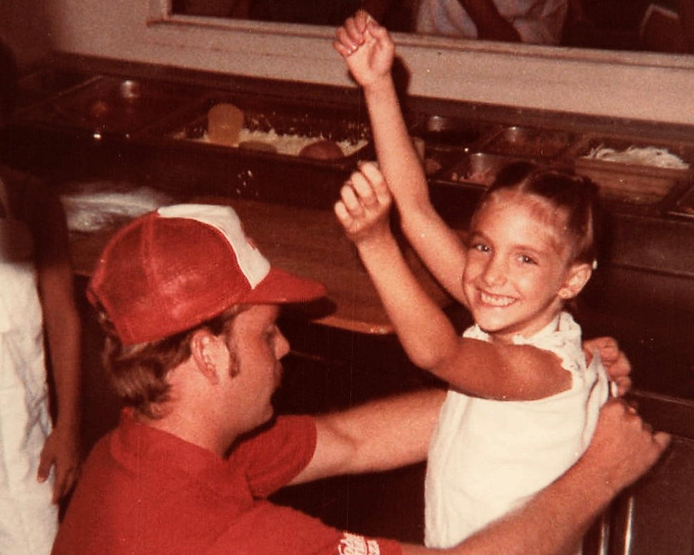 picture of founder cooking pizza at a young age. she's getting fitted with an apron with her arms up in the air smiling at the camera.